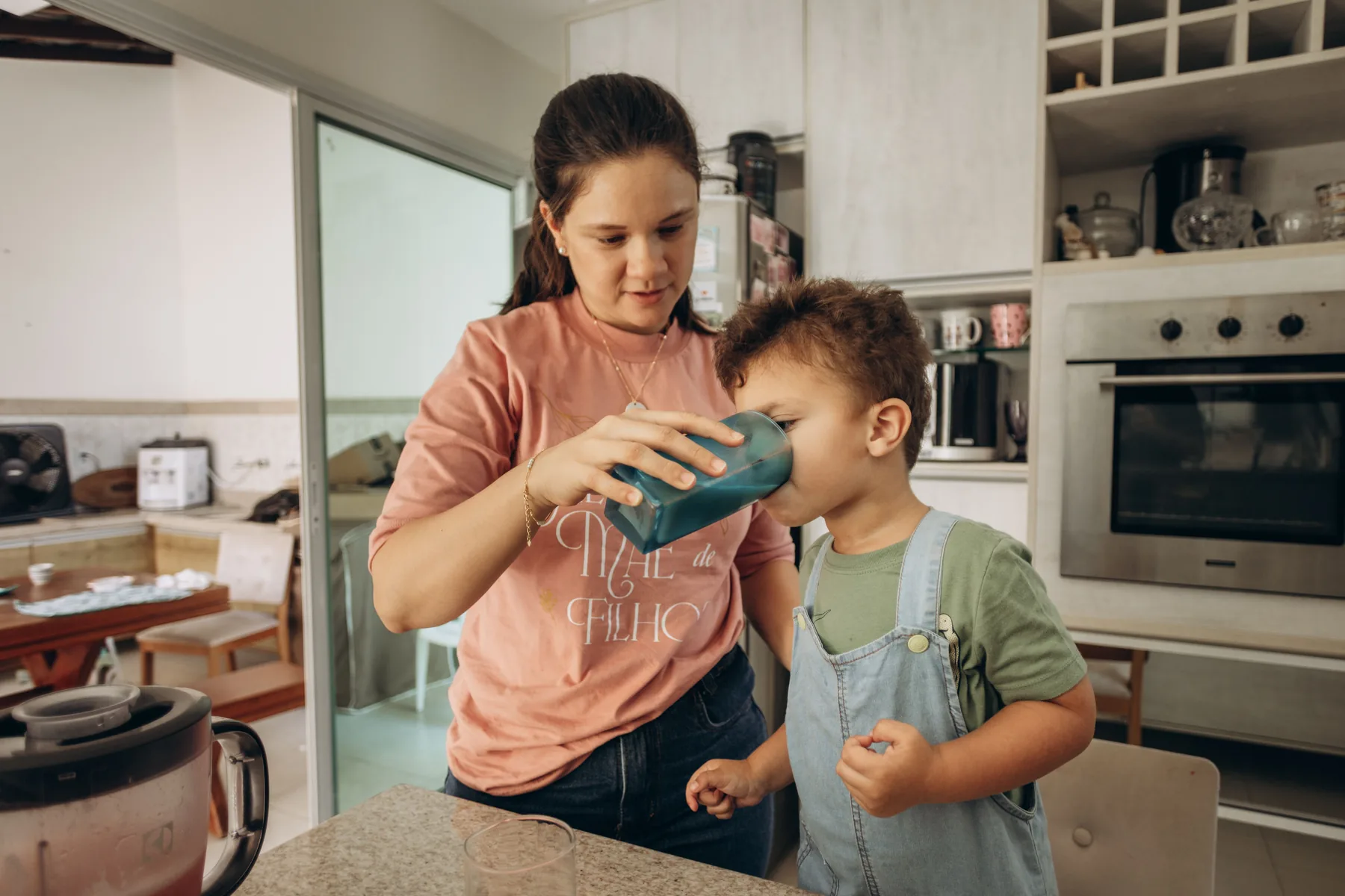 Parent helping a child drink water in a home kitchen